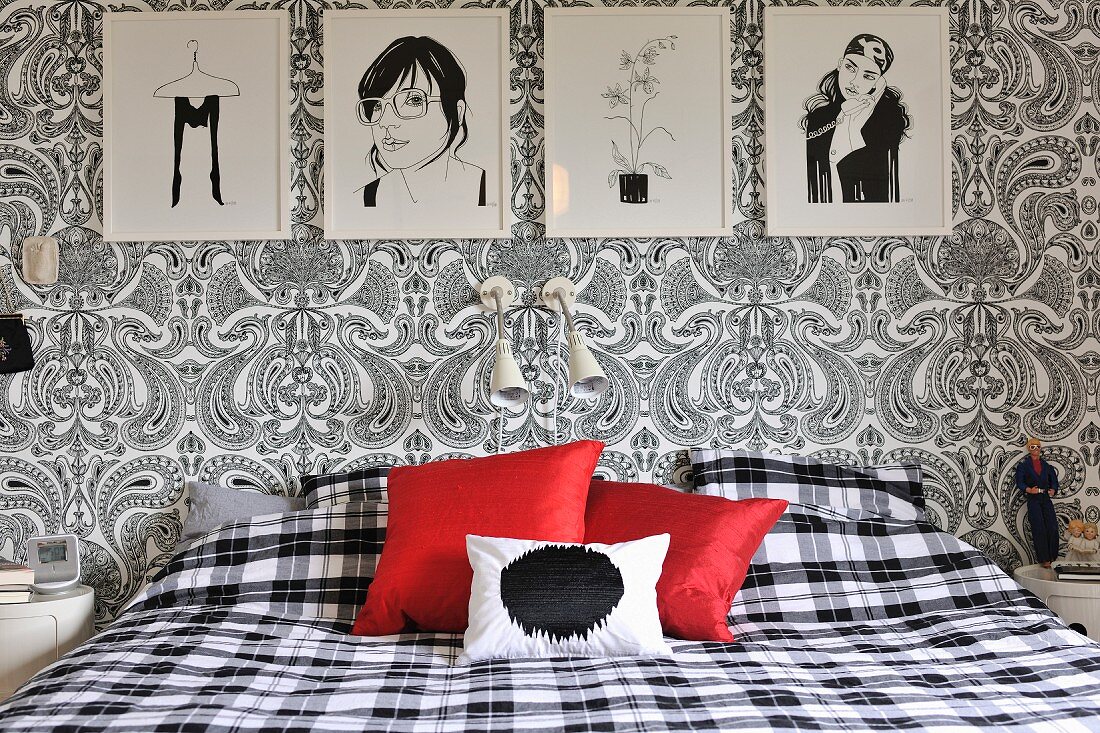 Bedroom with black and white drawings, floral Paisley wallpaper, gingham bed linen and red scatter cushions providing a splash of colour