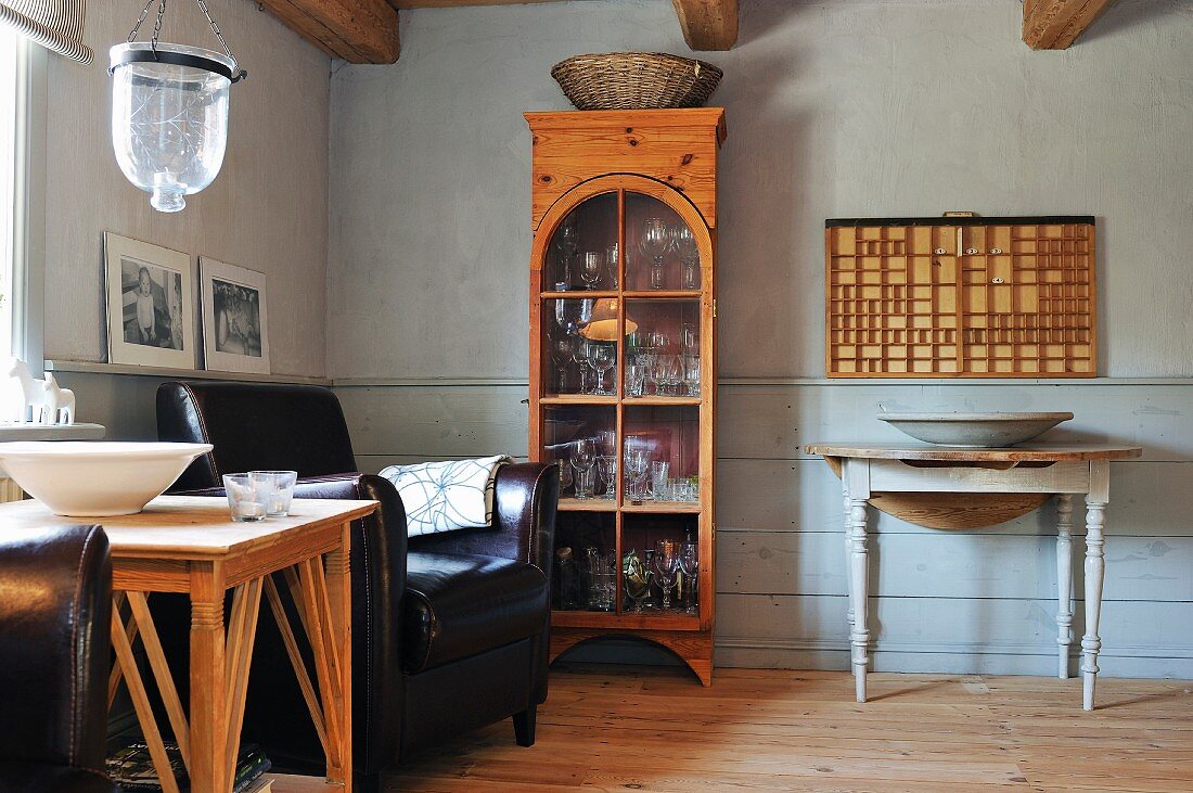 Landhausambiente mit Vitrinenschrank, Ledersessel und Holztisch mit gedrechselten Tischbeinen vor hellgrauer Wandverkleidung