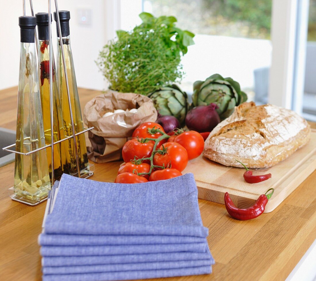 Serviettenstapel und Ölflaschen neben frischem Gemüse und Brot auf einer Kücheninsel mit heller Holzarbeitsplatte
