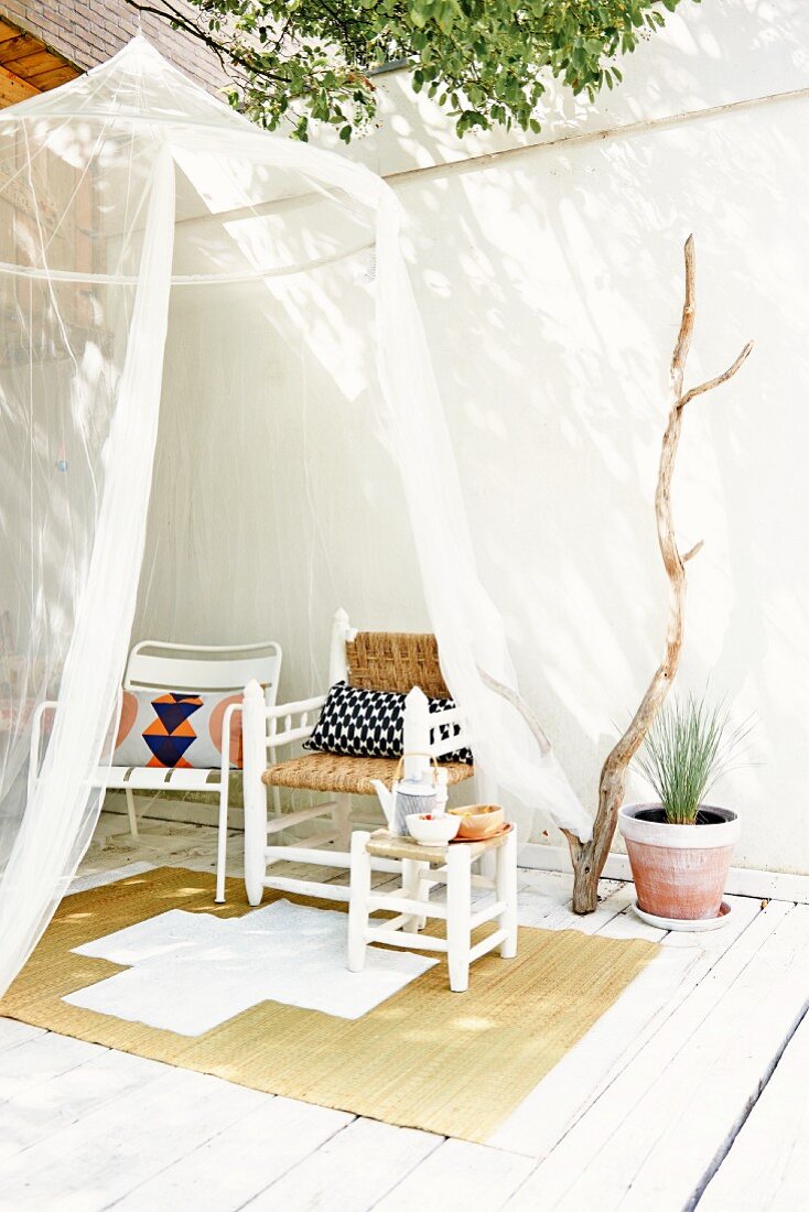 Moskitonetz aus transparentem Stoff auf Terrasse am Baum aufgehängt, Hocker und verschiedene Stühle auf Basttepich und weiss gestrichenem Holzboden