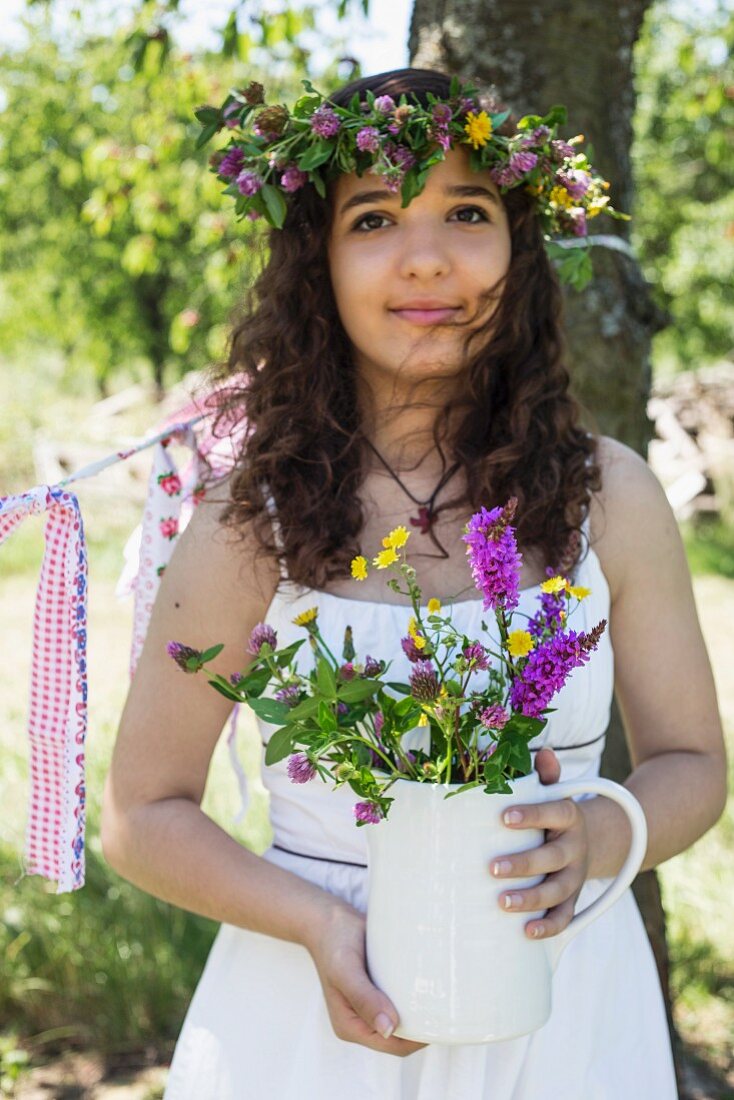 Junges Mädchen mit weißem Kleid und Blumenkranz auf Kopf, in den Händen Keramikkrug mit Wiesenblumen