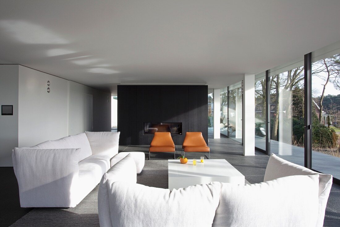weiße Polstersofagarnitur auf dunkelgrauem Boden in offenem, minimalistischem Wohnraum, im Hintergrund schwarzer Raumteiler, seitlich verglaste Fassade mit Panoramablick