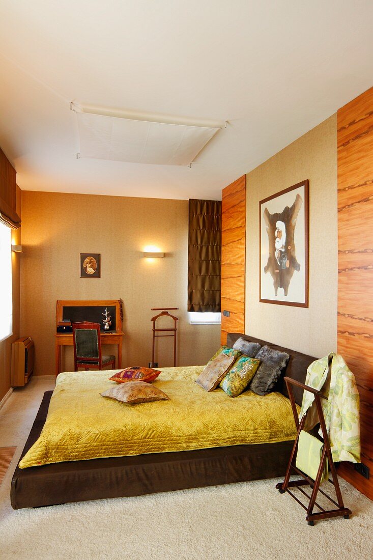 Niedriges Bett mit glänzender, gelber Tagesdecke vor holzverkleideter Wand, im Hintergrund Stuhl und Tisch an beleuchteter Wand