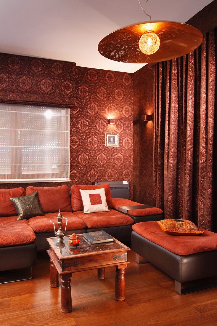 Sofakombination in Rostrot und folkloristischer Couchtisch in Lounge mit gemusterter Tapete und passendem Vorhang, an Decke Hängeleuchte mit Metallschirm