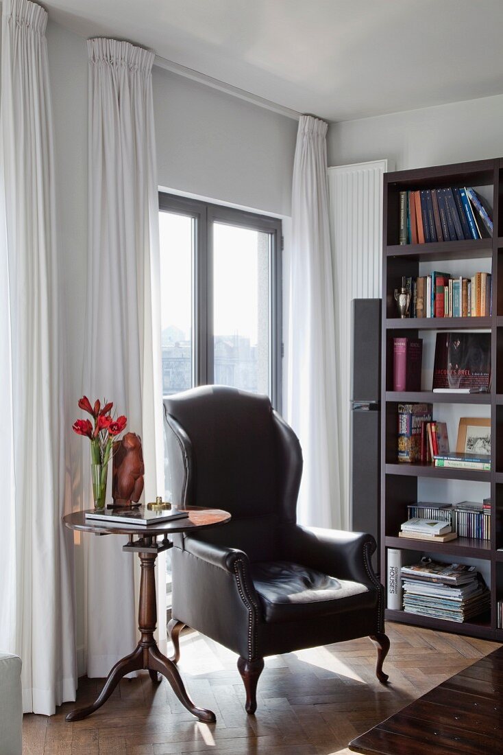 Antique armchair with leather cover and side table in front of balcony doors with modern bookcase to one side