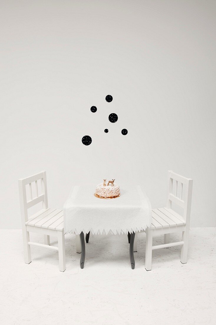 Tischdecke mit Zackenrand und mit Tierfiguren verzierter Kuchen auf Kindertisch; dahinter schwarze Punkte als Wanddeko