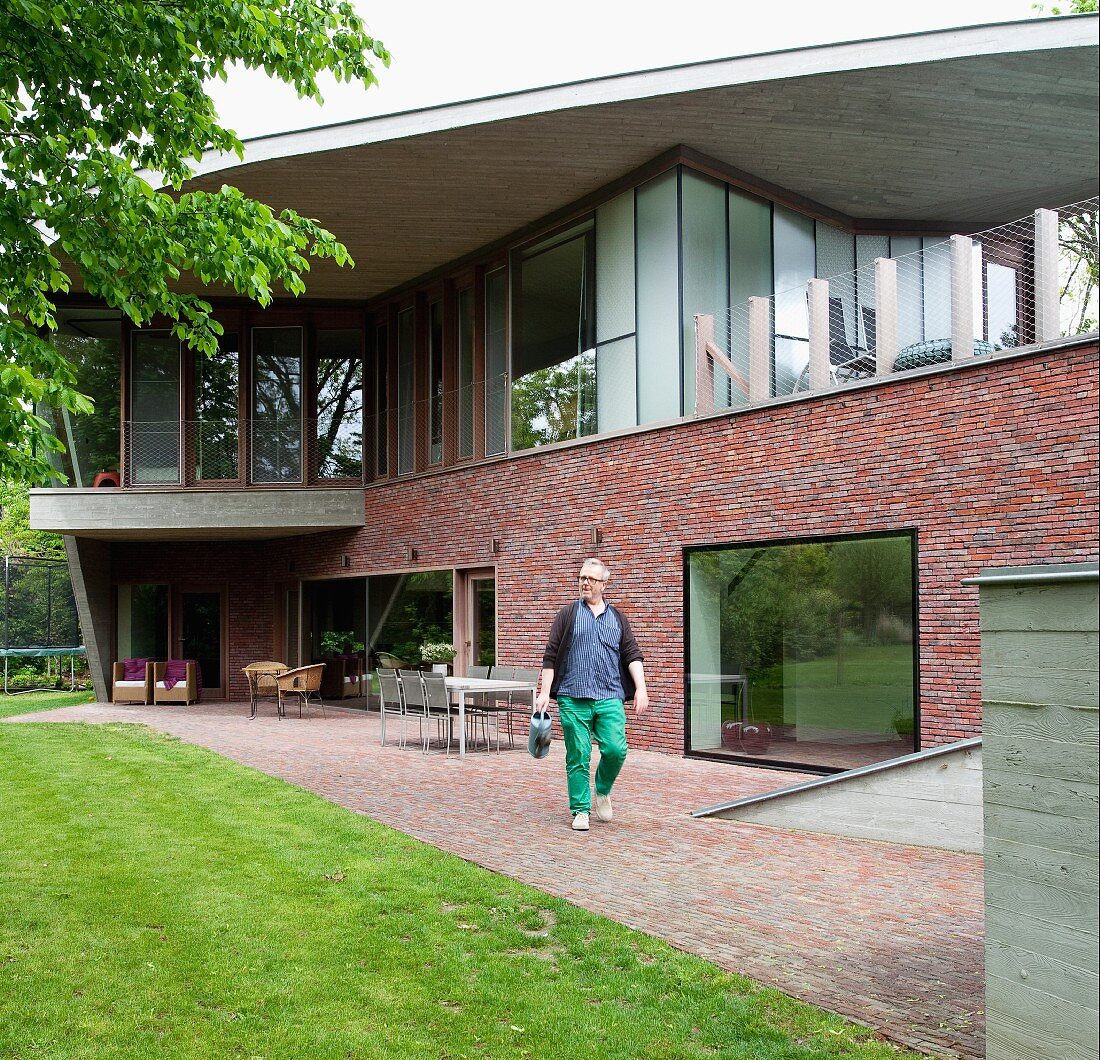 Terrace in back garden of Belgian, architect-designed house with brick facade, large windows and protruding concrete roof; man with watering can in foreground