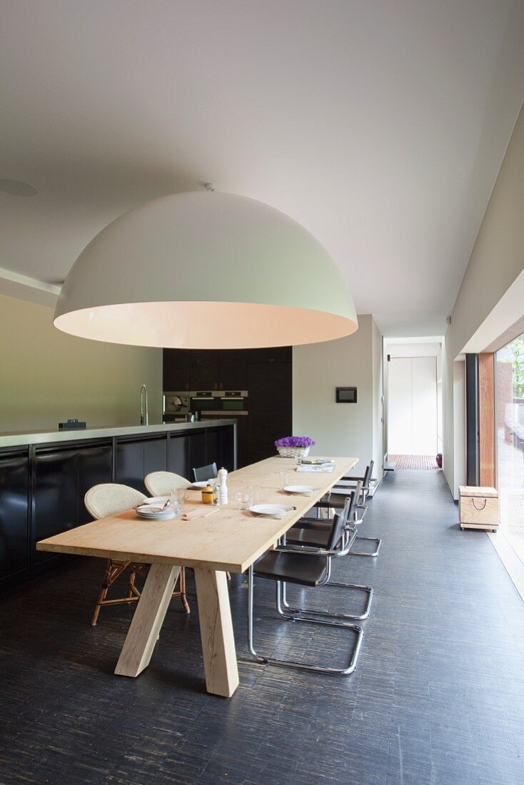 Long, pale wooden table and classic cantilever chairs below gigantic hemispherical lamp; kitchen base units with black doors