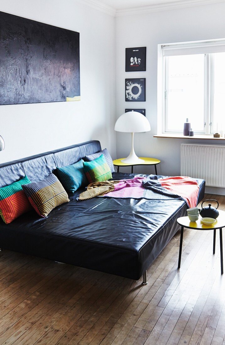 Large, black leather sofa with colourful scatter cushions and blanket, small, yellow retro table and white table lamp in living room