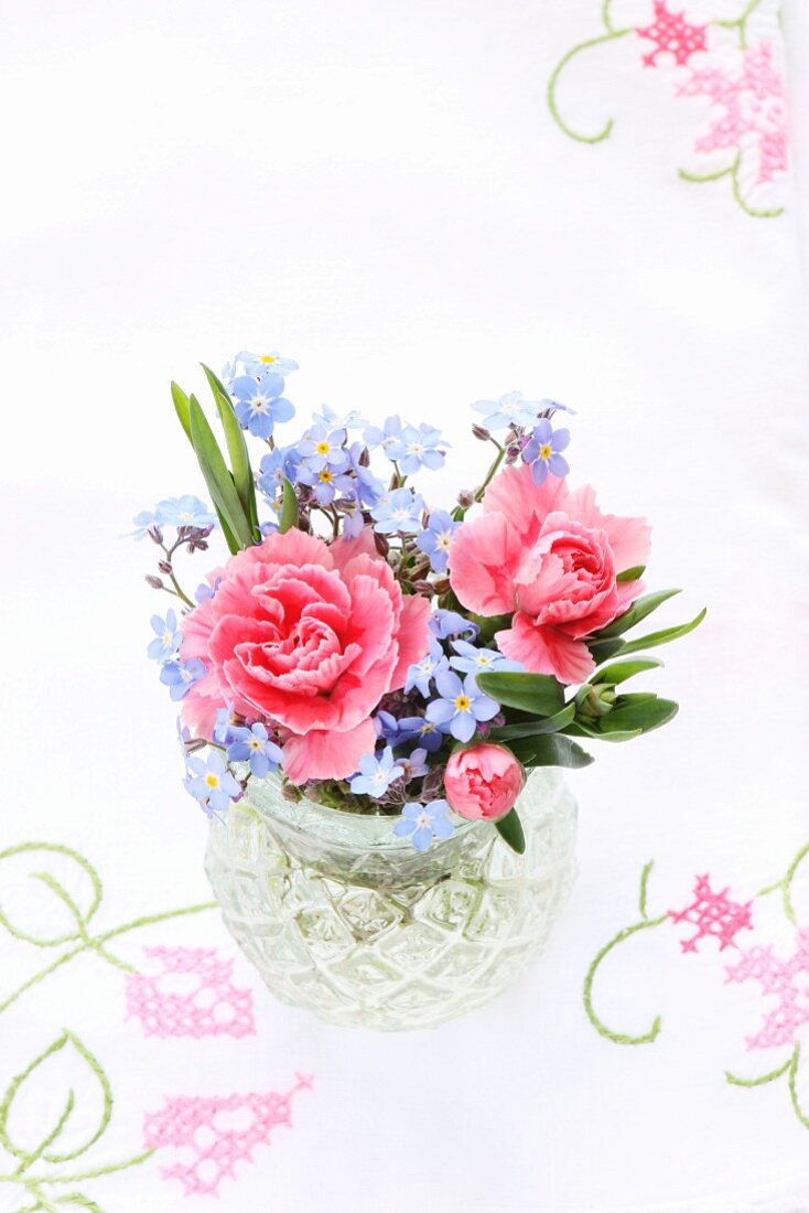 Vase mit Blumenstrauss aus rosa Nelken und Vergissmeinnicht auf bestickter Tischdecke