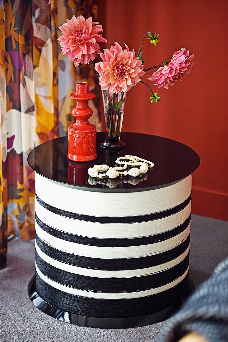 Dahlienstrauss und ziegelrote Keramikvase auf rundem, schwarzweiss gestreiftem Coffeetable; floraler Vorhang im Hintergrund