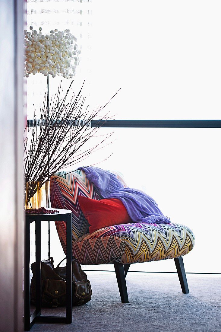 Rotes Kissen auf Sessel mit bunt gestreiftem Zickzackmuster, daneben Zweige in Vase auf Beistelltisch und Designerlampe