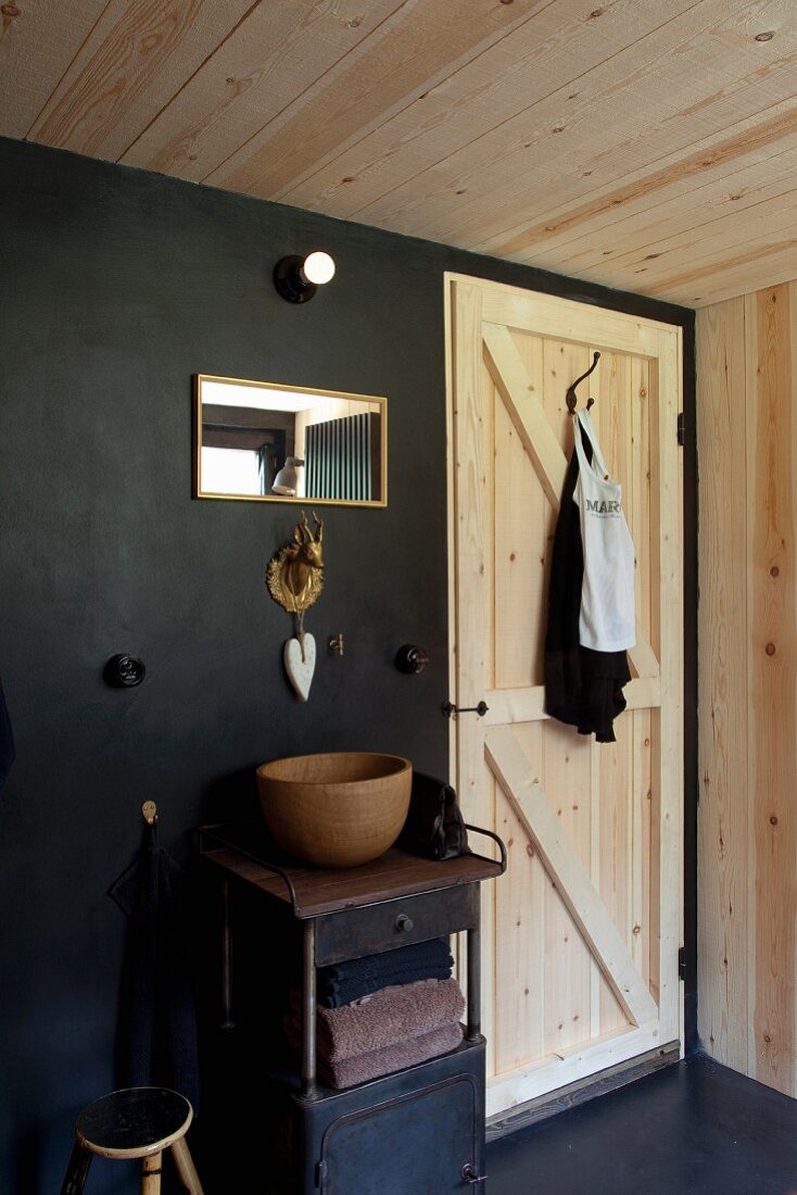 Wooden washbasin on small, vintage metal cabinet with brass stag's head as tap fitting on black wall next to plain wooden door