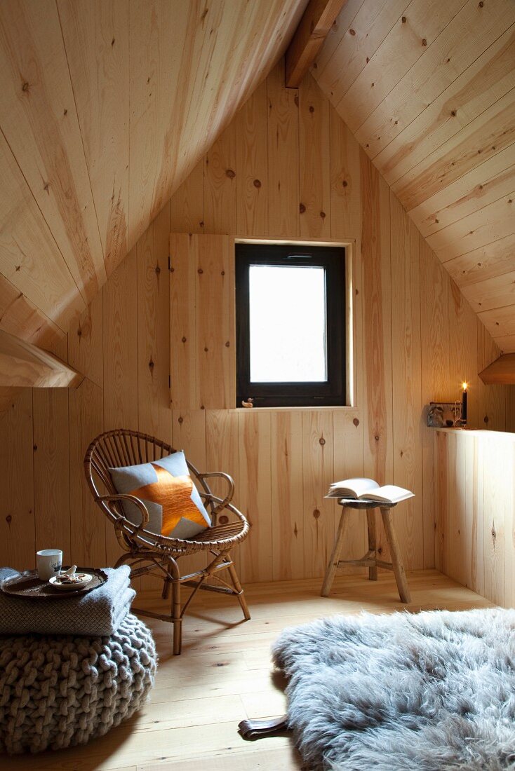 Holzverkleideter Rückzugsort unter Satteldach, Rattanstuhl mit Kissen und gemütlicher Sitzpouf auf Holzboden mit Schaffell