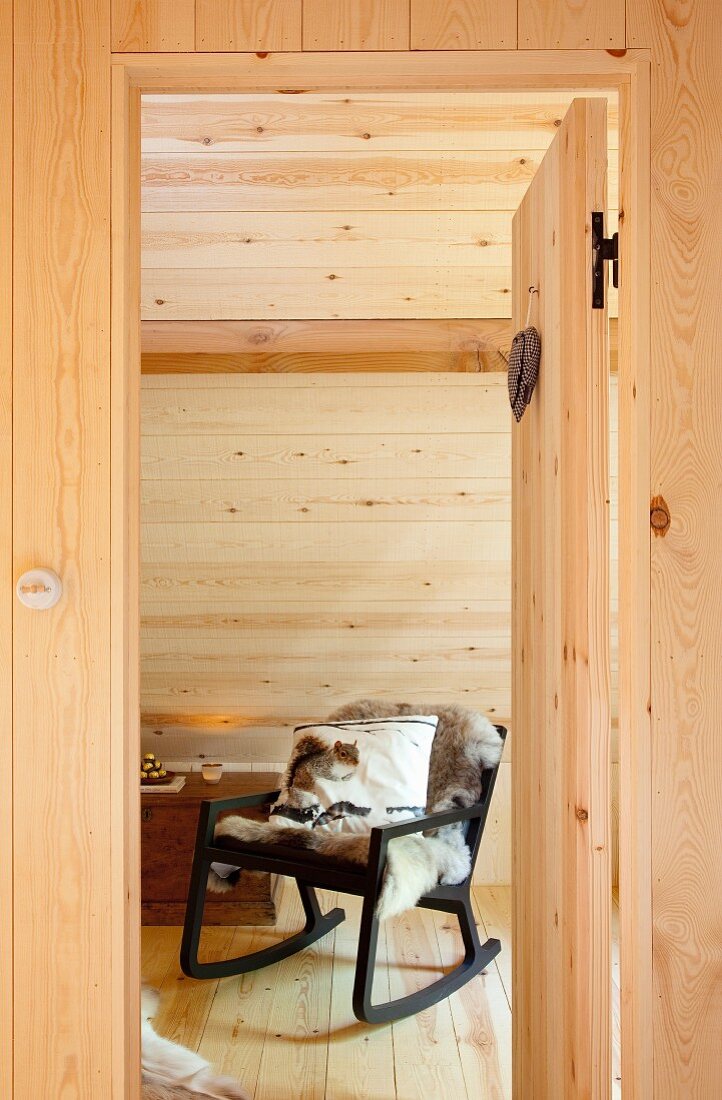 View through open interior door of comfortable, black rocking chair with animal-skin blanket and scatter cushion below sloping ceiling