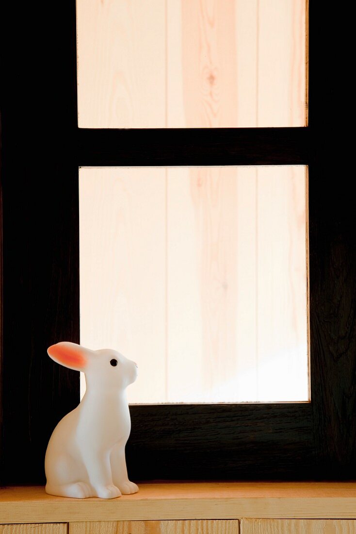 weiße Hasenfigur auf Holzfenstersims vor schwarzem Fensterrahmen