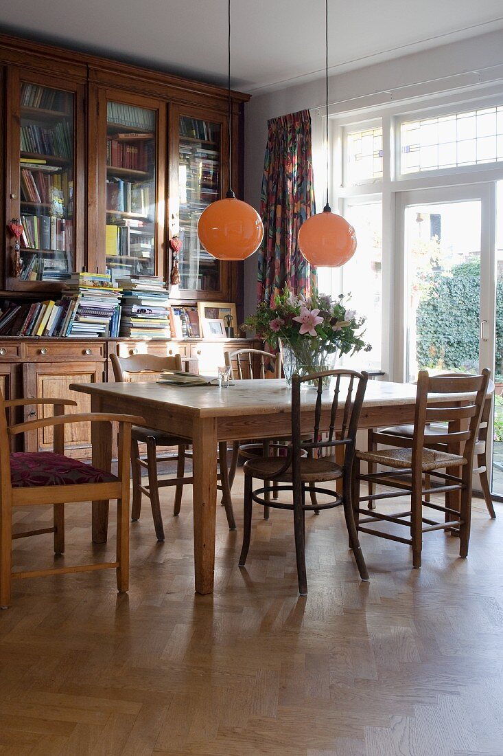 Dining area with wooden table and various wooden chairs below spherical pendant lamps in corner of living room