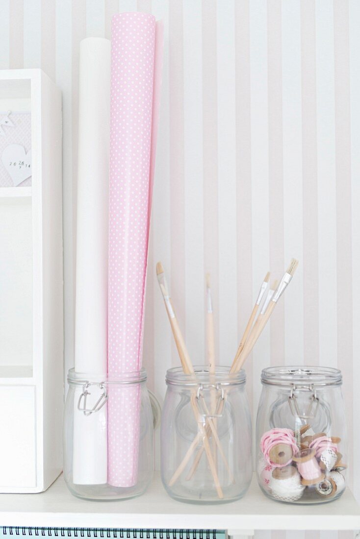 Storage jars holding pretty paper, paintbrushes and ribbons against pale, striped wallpaper