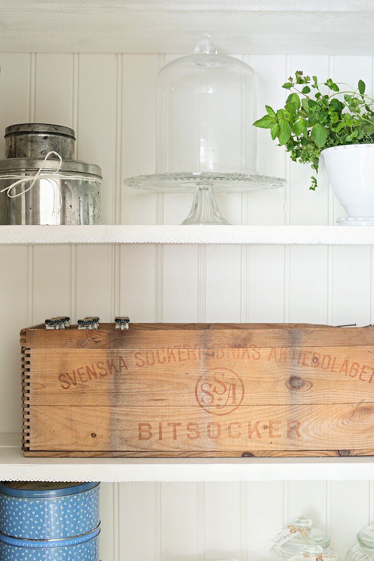 Vintage Holzkiste mit schwedischem Aufdruck, Blechdosen und Etagere mit Glashaube auf Regalborden