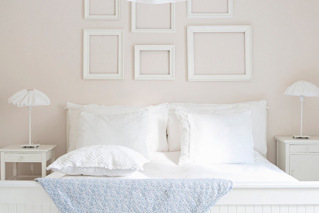 Weiß bezogenes Doppelbett, flankiert von Nachtkästchen mit Rüschenleuchten, an der Wand kassettenartig angeordnete, leere Bilderrahmen