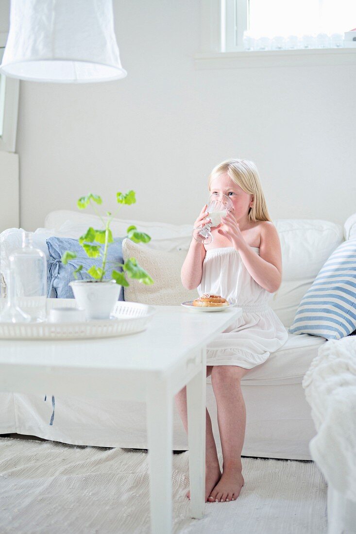 Blondes Mädchen trinkt Milch auf weißem Sofa