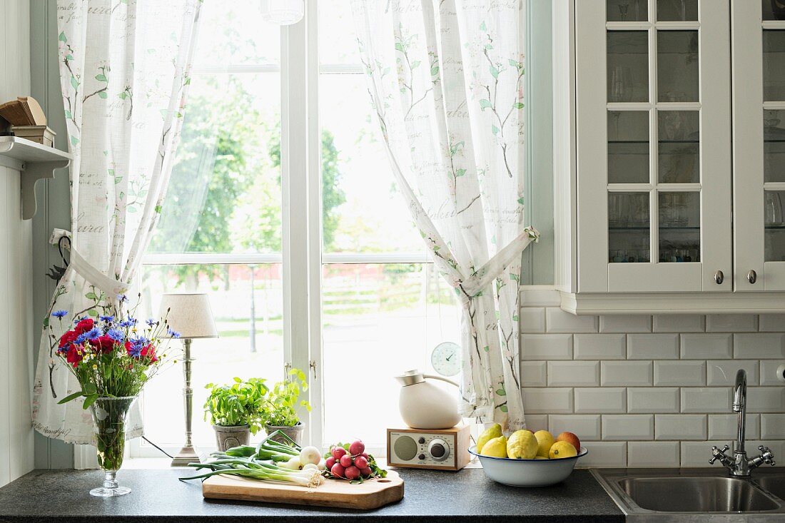 Schneidebrett mit Gemüse, seitlich Blumenstrauss in Vase auf Küchenzeile vor Fenster mit gerafftem Vorhang