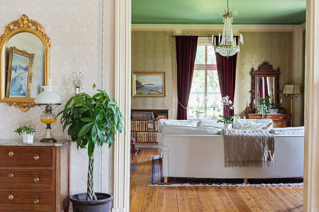 Vorraum mit Zimmerbäumchen neben breitem Durchgang und Blick auf helle Couch in traditionellem Wohnzimmer, Kronleuchter an grüner Decke