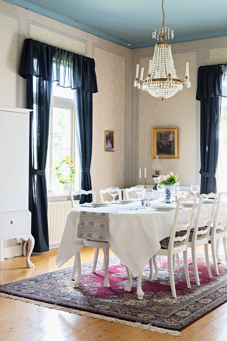 White chairs with carved backrests around festively set table below chandelier in traditional dining room with windows