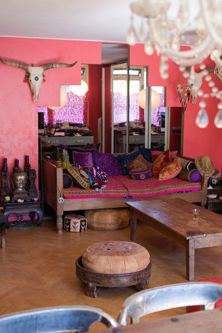 Eklektizistischer Stil; gemütliches Wohnzimmer mit gepolsterter Sofabank und vielen Kissen, abgetrepptem Wandspiegel, Tiertrophäe und rosafarbener Wand
