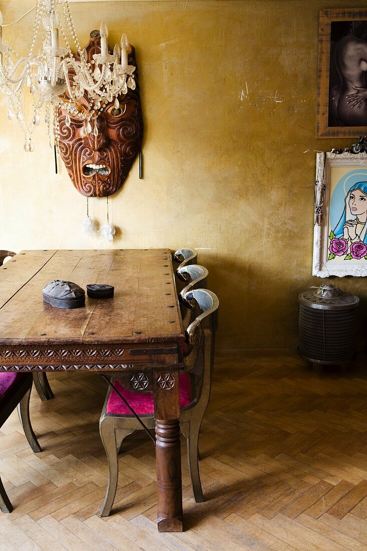 Kunsthandwerkliche asiatische Holzmaske an goldfarbener Wand, Kronleuchter und Edelholztisch mit Schnitzereien in eklektischem Ambiente
