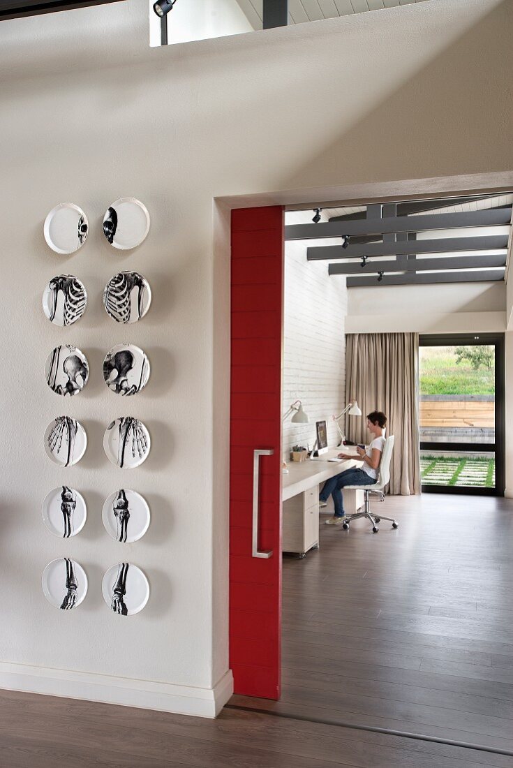 Wandteller mit Anatomie-Design neben offenem Durchgang mit roter Schiebetür und Blick auf Home Office