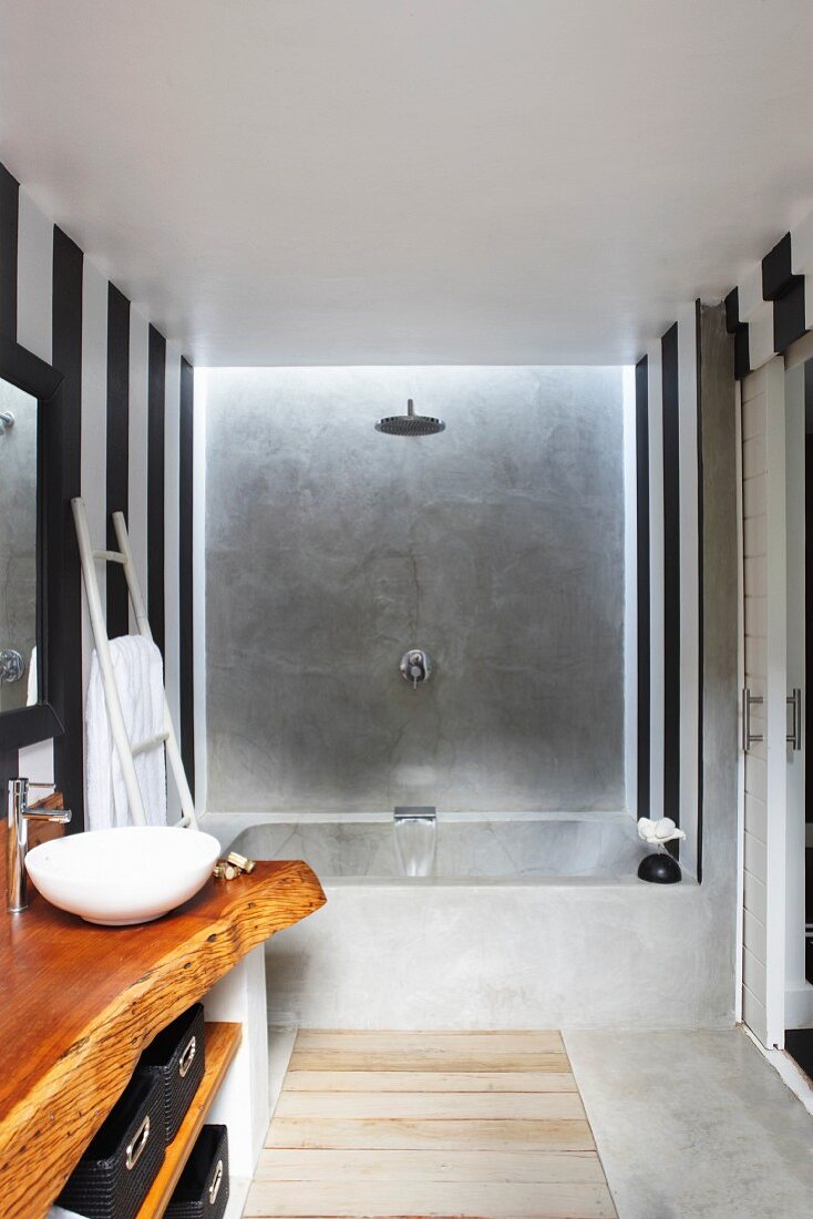 Designerbad, seitlich Waschtisch mit rustikaler Holzplatte, vor Wand mit schwarz weissen, vertikalen Streifen, im Hintergrund Badewanne aus Beton