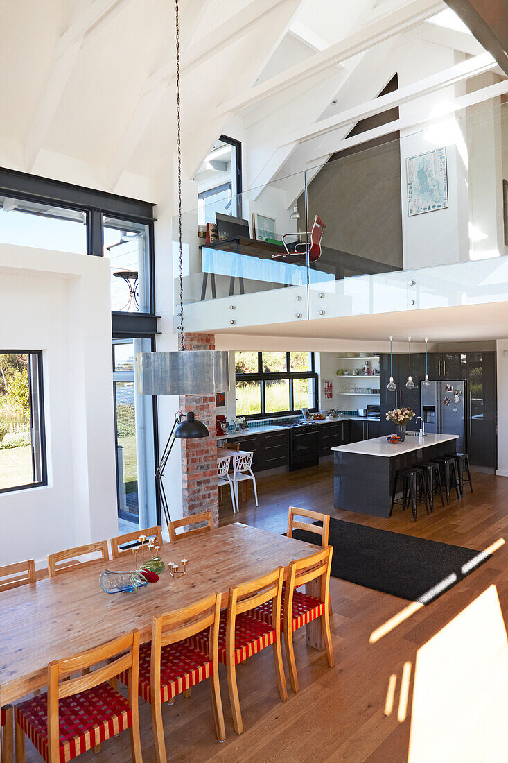 Dining area with chairs and long wooden table and kitchen below gallery in background in open-plan, modern interior