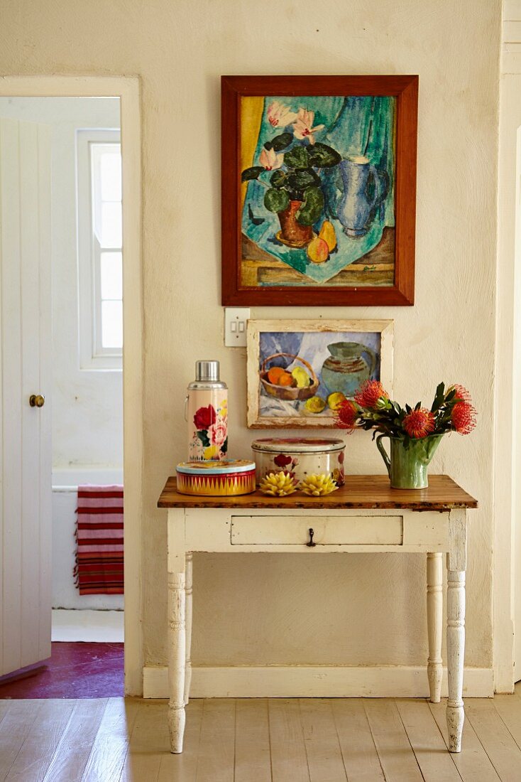 Vintage containers and vase of proteas on rustic console table below framed pictures in foyer