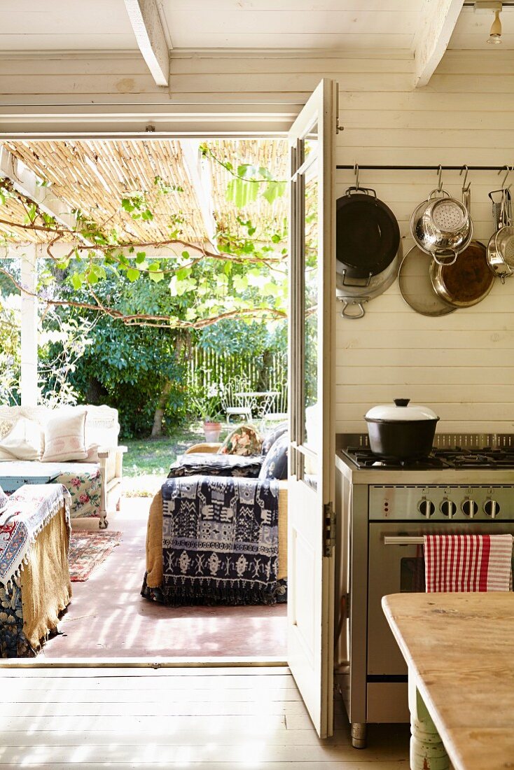 Blick von Küche durch offene Tür auf überdachte Terrasse mit gemütlichen Polstersofas, vor sommerlichem Garten