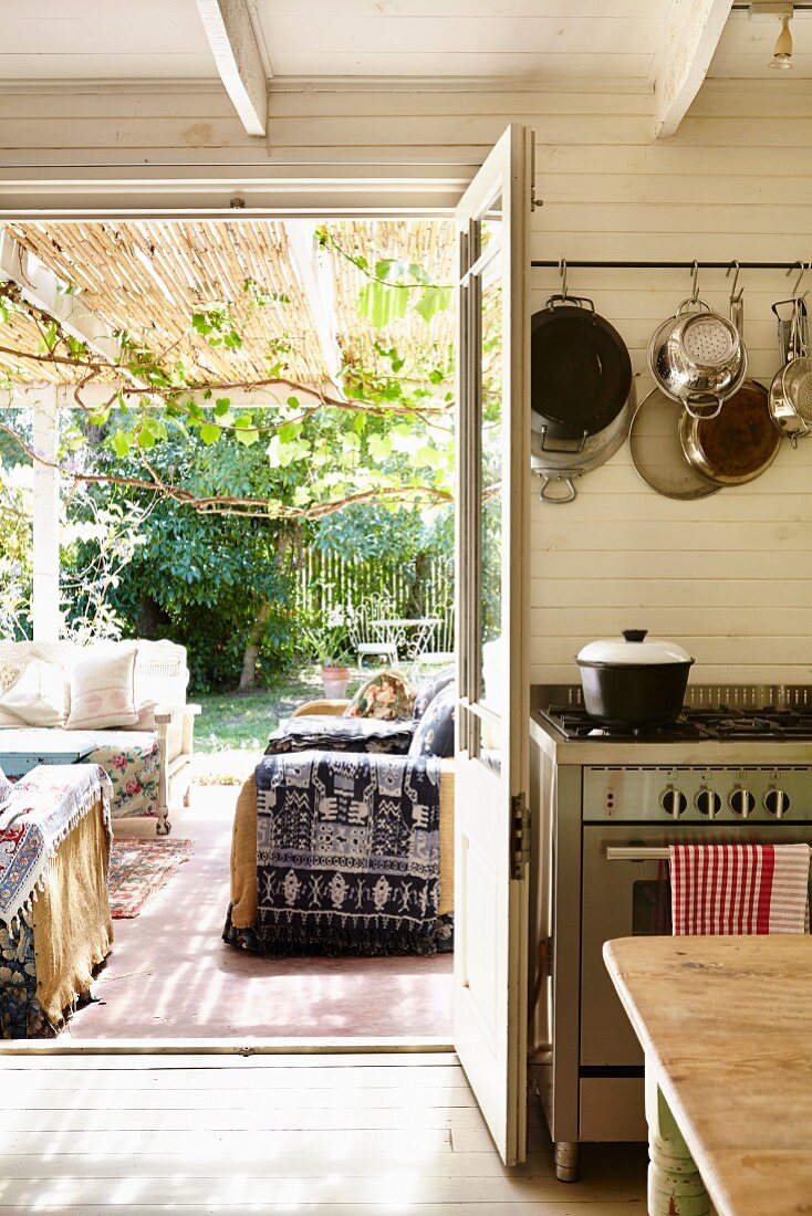 View from kitchen through open door onto roofed terrace with comfortable sofas and summery garden