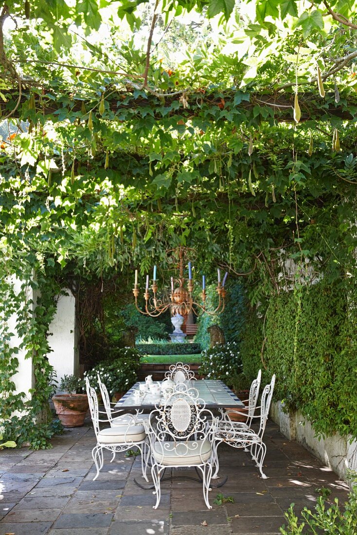 Ornate, white metal chairs around table on terrace and candle chandelier hanging from pergola