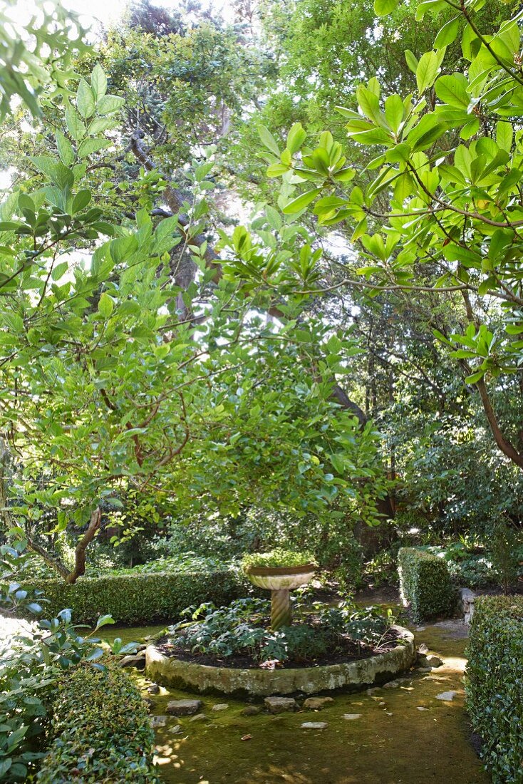 Rundes Beet mit Steineinfassung, in der Mitte Pflanzgefäss auf Stele, in dicht bewachsenem Garten