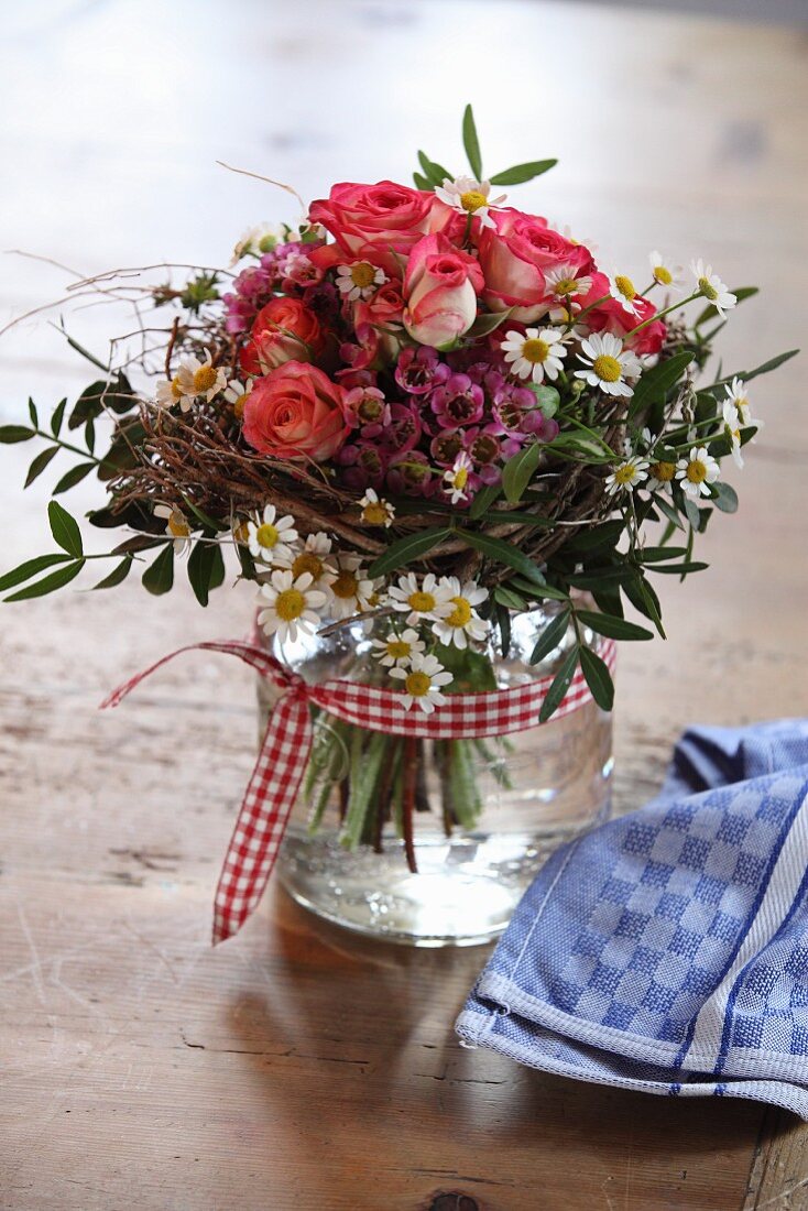 Blumenstrauss aus Rosen und Gänseblümchen im Glas, mit kariertem Schleifenband verziert
