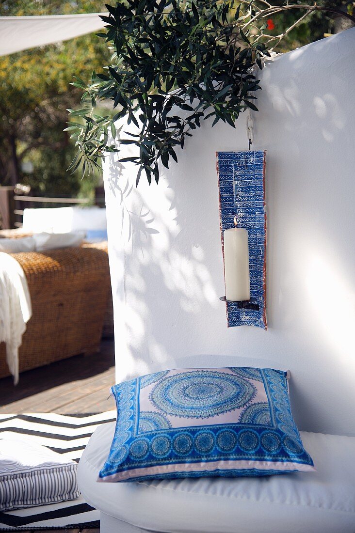 Blau-weiss gemustertes Kissen auf Sitzbank, vor Rückwand mit aufgehängtem Kerzenhalter und Kerze auf mediterraner Terrasse