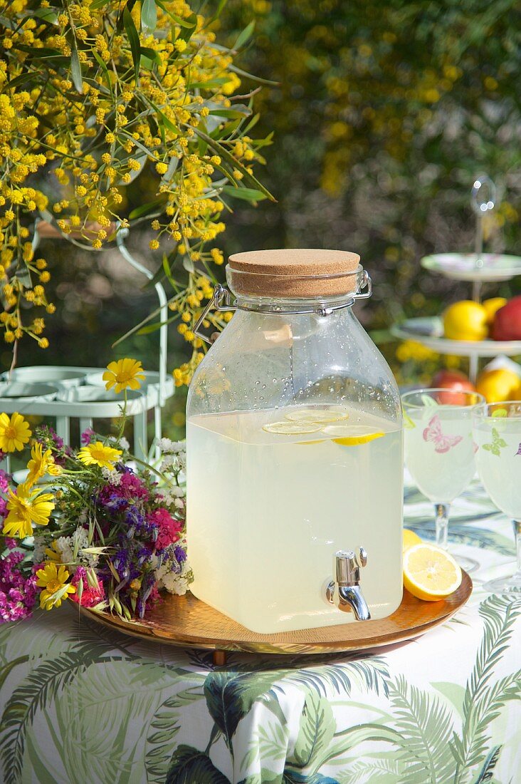 Selbstgemachte Zitronenlimonade in Vintage-Glasbehälter auf Tablett mit buntem Wiesenblumenstrauß im Freien
