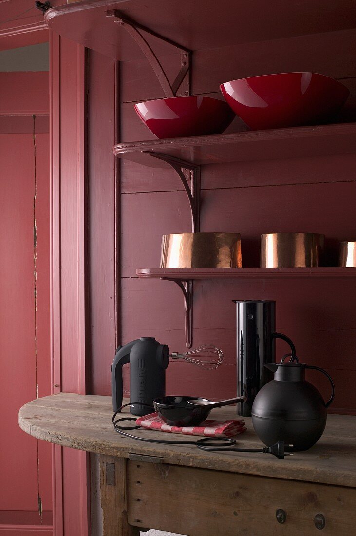 Verschiedene Schüssel auf Wandregalen und Küchengeräte auf Holztisch in dunkelroter Vintage Küche
