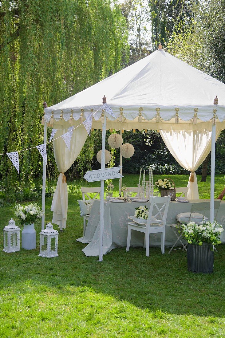 Hochzeitsfeier im Garten, weisses Zeltpavillon mit Girlanden dekoriert, davor Bodenlaternen und Blumensträusse in Gefässen auf der Wiese