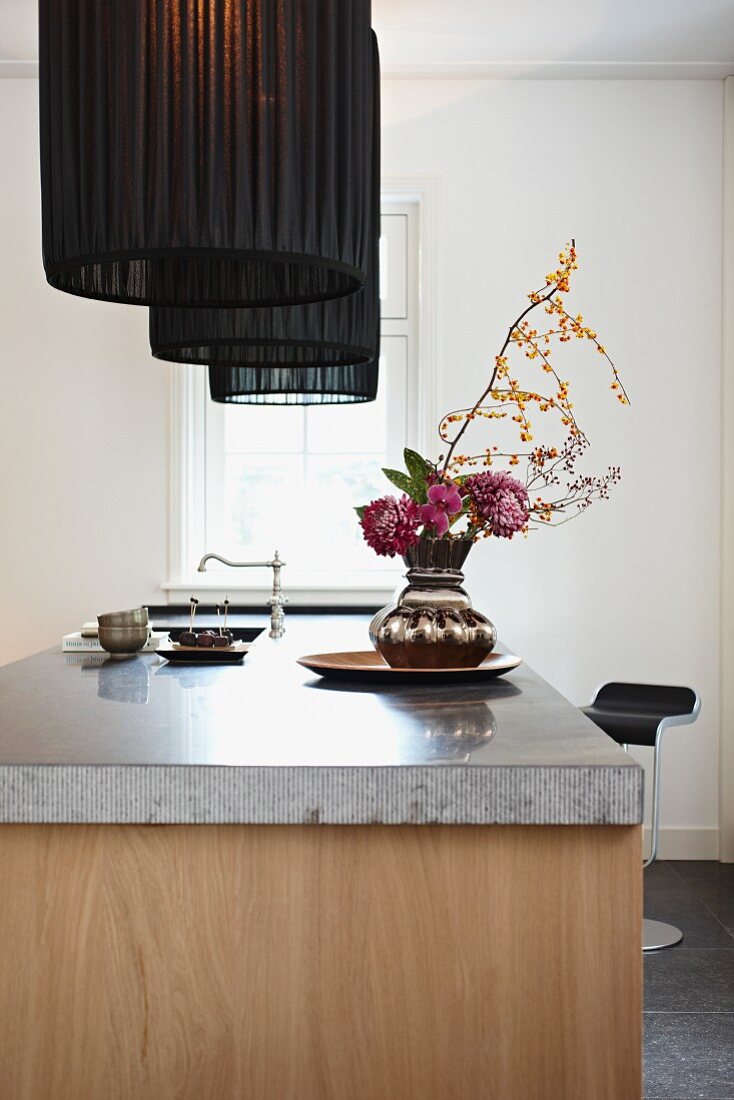 Bouquet in silver vase on free-standing kitchen counter with stone worksurface below pendant lamps with black fabric lampshades