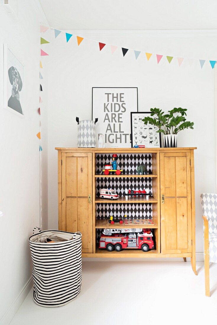 Toys on open-fronted shelves of wooden sideboard and black and white striped basket in corner of child's bedroom with colourful bunting on wall