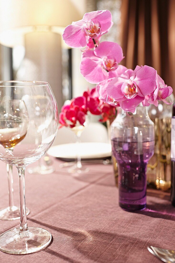 Pinkfarbene Orchideenrispen in Glasvase mit farbigem Wasser und Weingläser auf gedecktem Tisch