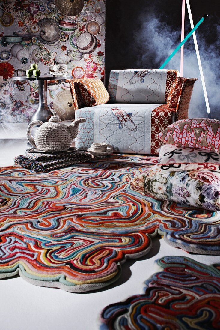 Künstlerisch gestalteter, bunter Teppich, gemusterte Kissen, gehäkelte, stilisierte Teekanne und Tasse vor Sessel