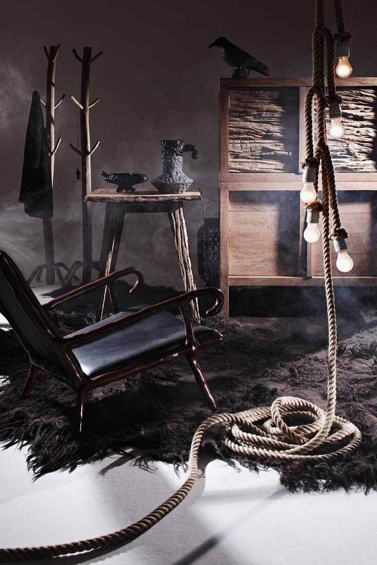 Geheimnisvolle Inszenierung im archaischen Naturlook - schwarzer Ledersessel mit knorzigem Holzgestell auf Zottelteppich und Hängeleuchte in Seil-Optik