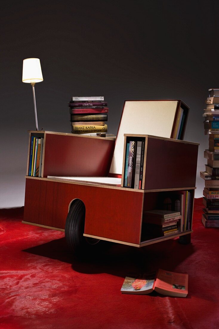 Multifunctional armchair made from laminated wood with large wheel, shelf openings for books and table lamp on red carpet