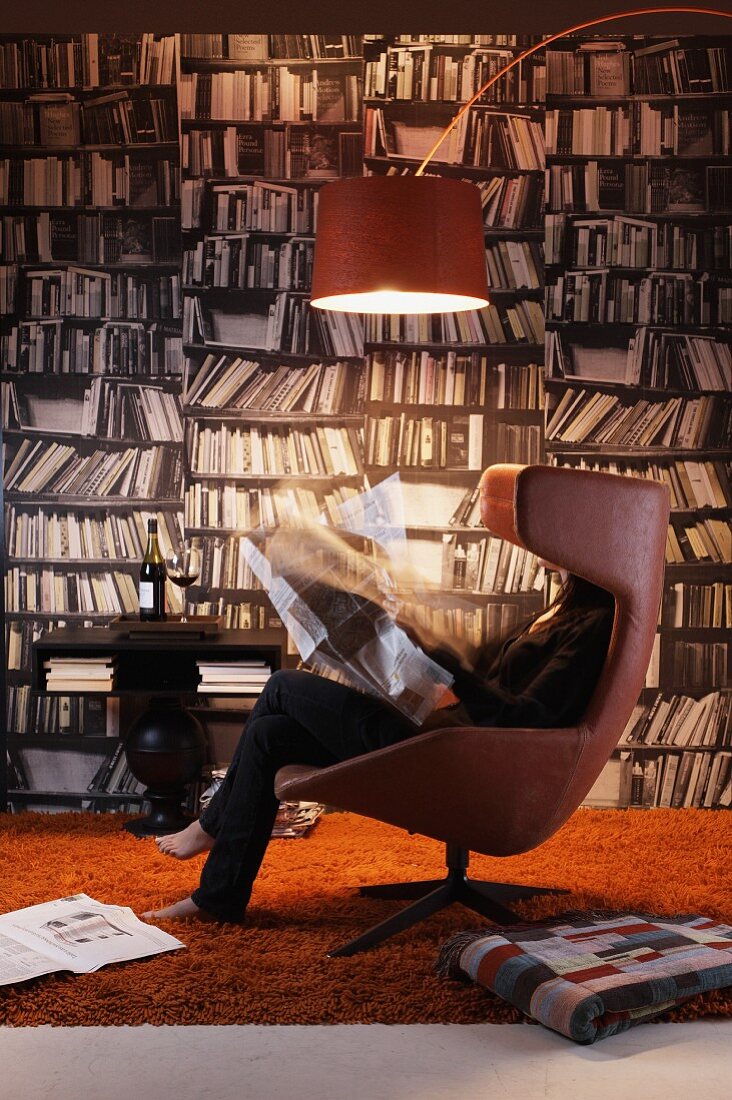 Woman reading in designer, leather easy chair below arc lamp in front of photo mural of bookcase